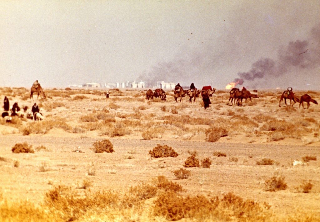 Colour photograph of camels and herdsmen walking in scrubland, while tall buildings and a burning oil well are visible in the distance